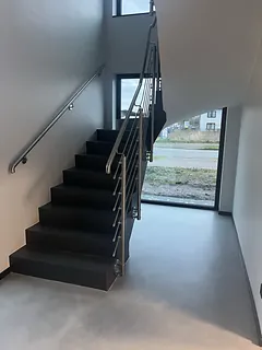 Treppenhaus im Erdgeschoss mit fugenlosem grauem Boden, dunkler Treppe, Edelstahlgeländer und bodentiefem Fenster